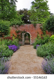 An English Walled Garden In Early Summer With Path Leading To An Arched Gate