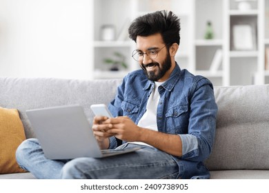 Engrossed indian man in eyeglasses multitasking with laptop and smartphone at home, young eastern freelancer guy messaging on mobile phone and using computer while sitting on couch at home - Powered by Shutterstock