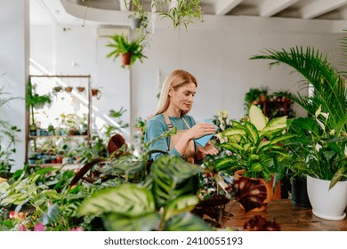 Engrossed Middle-Aged Female Businesswoman, Clad in Apron, Methodically Dusting the Leaves of a For-Sale Plant in her Flower Shop, Leading a Floristry Workshop Amidst the Serene Greenery. - Powered by Shutterstock