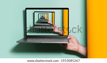 Enigmatic surrealistic optical illusion. Close-up of male hand holding laptop on blue and orange background.