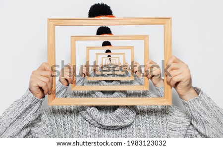 Enigmatic surrealistic optical illusion; portrait of young man holding frame on grey background.
