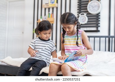 Enjoin sister teaching little boy brother to read a book together while sitting on the bed at home. - Shutterstock ID 2250537251