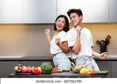 Enjoy Cooking. Joyful Asian Couple Dancing Preparing Dinner And Having Fun In Kitchen At Home. Happy Family Weekend Leisure, Food Preparation And Recipes, Nutrition Concept. Empty Space For Text