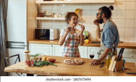 Enjoy it. Young man and woman in apron eating freshly baked pizza and drinking wine while standing in the kitchen. Love, relationships concept. Web Banner - Powered by Shutterstock