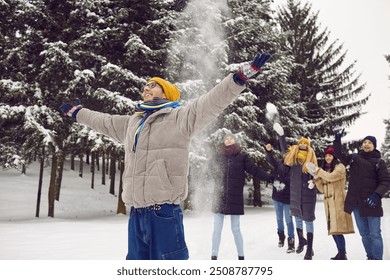 Enjoy little things. Cheerful and carefree young man stands with arms outstretched against background of friends throwing snowballs at him. Friends spend time together and have fun in snowy forest. - Powered by Shutterstock