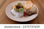 enjoy Nasi uduk with a side dish of martabak, crackers, egg slices and cucumber slices served on a round plate on a wooden table.