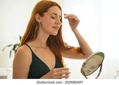 Enjoyable red hair young girl doing beauty routine with facial moisturizer at home. Redhead smiley beautiful woman using natural cosmetic and looking to the mirror.
