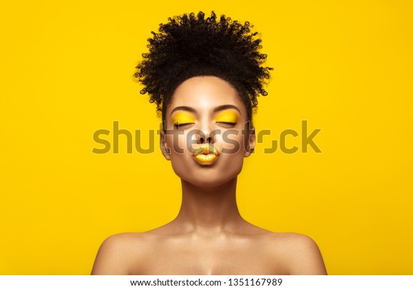 Enjoyed African American Fashion Model\
portrait . Satisfied Brunette young woman with afro hair style and\
closed eyes show kiss, creative yellow make up, lips and eyeshadows\
on colorful background.