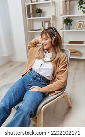Enjoyed Free Time Young Blonde Student Lady Freelancer In Warm Sweater In Headphones Listen Fav Songs Sitting In Armchair At Modern Home Interior. Music Time Relaxing Cool Playlist Concept. Copy Space