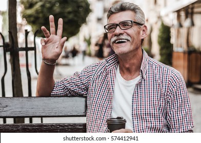 Enjoying a beautiful day. Cheerful mature man sitting on wooden bench with  a cup of coffee and smiling while waving to someone. - Powered by Shutterstock