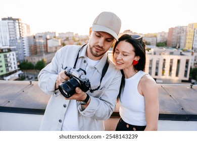 Enjoying beautiful sunset on rooftop, couple reviews photos on camera. Man, wearing cap and casual jacket, stand beside woman, smiles warmly, relaxed and happy expressions against cityscape background - Powered by Shutterstock