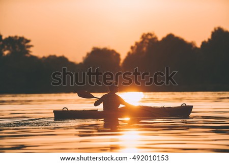 Enjoying the best sunset on river. Side view of young man kayaking on river with sunset in the background