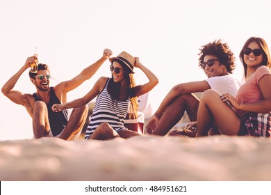 Enjoying carefree time with friends. Cheerful young people spending nice time together while sitting on the beach and drinking beer - Powered by Shutterstock