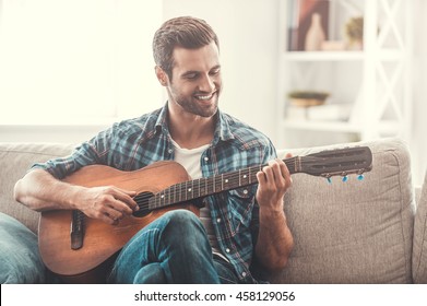 Enjoying carefree time at home. Happy young man playing the guitar while sitting on sofa at home