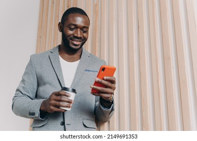 Enjoying Coffee Break At Work. Young Happy Positive African Man Office Worker In Suit Using Mobile Phone And Chatting With Friends, Texting Sms Or Checking Email, Looking At Smartphone And Smiling