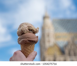 Enjoying a double scoop of gelato in Vienna, Austria. A hand holds a waffle cone with chocolate and vanilla ice cream, with St. Stephen's Cathedral blurred in the background against a blue sky. - Powered by Shutterstock