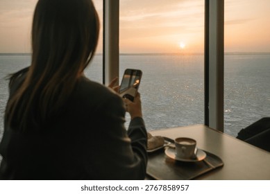 Enjoying a drink while using a smartphone to capture a sunset over the water
