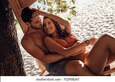 Enjoying every minute with each other.  Beautiful young couple embracing and looking at each other while resting on the beach together - Powered by Shutterstock