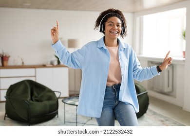 Enjoying Favorite Playlist. Excited Carefree Millennial African American Lady Listening To Popular Music In Wireless Headset Standing In Living Room And Dancing, Having Fun Looking At Camera