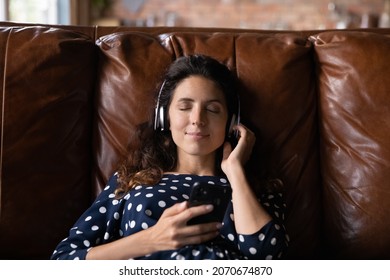 Enjoying favorite song. Calm latina woman in wireless headphones lie on sofa with closed eyes hold smartphone relieve stress by good tranquil music. Happy young lady listen to audio book in earphones