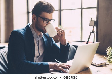 Enjoying Fresh Coffee At Work. Confident Young Man In Smart Casual Wear Working On Laptop And Drinking Some Hot Drink While Sitting At His Working Place In Office