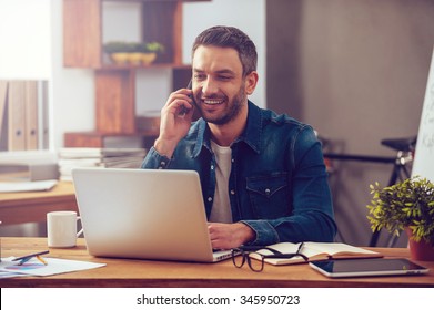 Enjoying good working day. Confident young man working on laptop and talking on the mobile phone while sitting at his working place in office - Powered by Shutterstock