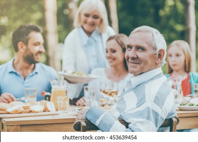 Enjoying great time with family. Happy family of five people sitting at the dining table outdoors while senior man looking over shoulder and smiling