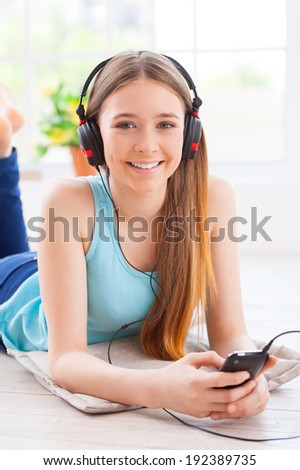 Enjoying her favorite music. Cute teenage girl in headphones listening to the music and looking at camera while lying down on the floor at her apartment