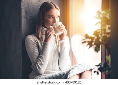 Enjoying her fresh coffee. Beautiful young woman drinking coffee and looking through window while sitting at windowsill at home