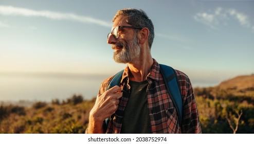 Enjoying the hilltop views. Happy mature hiker looking away with a smile on his face while standing on top of a hill with a backpack. Adventurous backpacker enjoying a hike at sunset. - Powered by Shutterstock