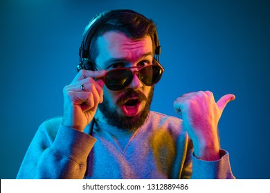 Enjoying his favorite music. Happy young stylish man in sunglasses with headphones listening and pointing by finger while standing against blue neon background