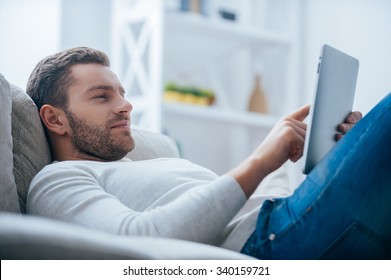 Enjoying his leisure time at home. Side view of handsome young man working on digital tablet and looking relaxed while lying on the couch at home - Powered by Shutterstock