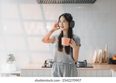 Enjoying moment of relaxation, young woman in cozy kitchen wears headphones while holding cup. bright space features modern appliances and minimalist design, creating cheerful vibe. - Powered by Shutterstock