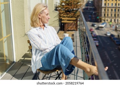Enjoying morning. Beautiful dreamy mature 60s woman in casual wear relaxing on balcony at home, starting new day with coffee. Happy elderly woman sitting on terrace and looking at sunny street