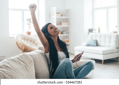 Enjoying music! Attractive young woman in headphones holding smart phone and gesturing while sitting on the couch at home