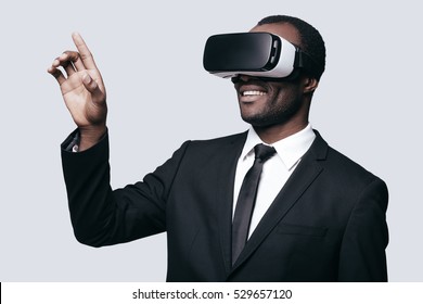 Enjoying new experience. Handsome young African man in VR headset gesturing and smiling while standing against grey background - Powered by Shutterstock