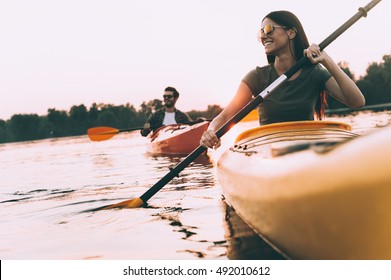 Enjoying nice time on river together. Low angle view of beautiful young couple kayaking on river together and smiling with sunset in the background