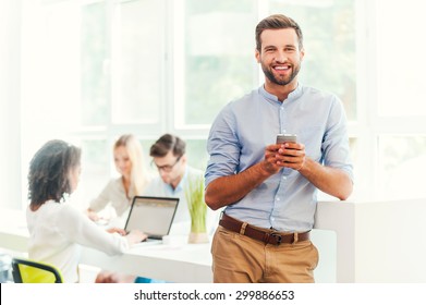 Enjoying Office Life. Joyful Young Man Holding Mobile Phone And Looking At Camera While His Colleagues Working In The Background