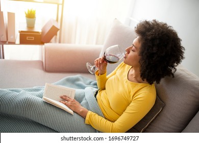 Enjoying a relaxing afternoon with a good book. Beautiful young African woman enjoying a glass of wine while relaxing at home. Beautiful young woman reading book near window at home.