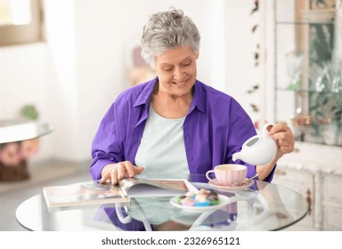 Enjoying Senior Years. Smiling Mature Woman Pouring Tea In Cup, Enjoys Drink And Reads Magazine On Weekend, Sitting At Table At Cafe Indoor. Gray Lady Tasting Drink And Desserts In Restaurant - Powered by Shutterstock