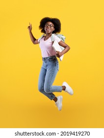 Enjoying student life. Full length portrait of african american lady jumping, wearing backpack and holding notepads over yellow studio background, crop
