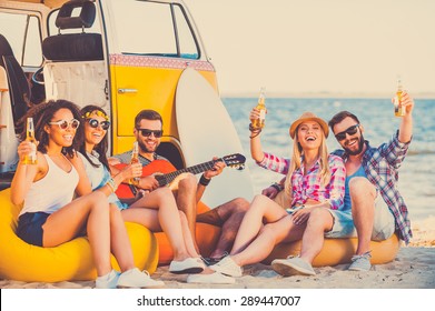 Enjoying summer time together. Group of happy young people having fun together while sitting on the beach near their retro van - Powered by Shutterstock