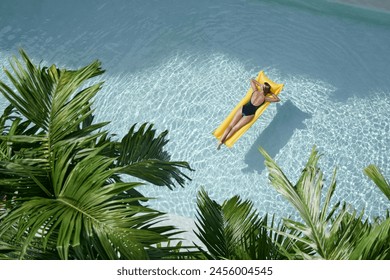 Enjoying suntan. Tropical vacation concept. Top view of young woman on the yellow air mattress in the swimming pool with palm trees. - Powered by Shutterstock