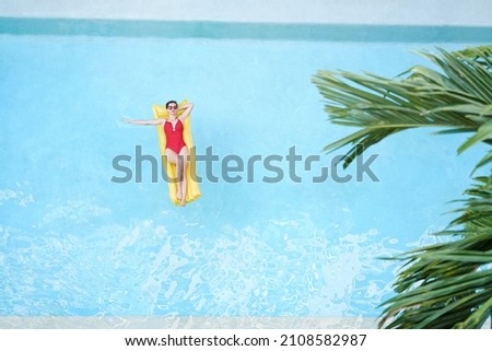 Enjoying suntan. Tropical vacation concept. Top view of young woman on the yellow air mattress in the swimming pool with palm trees.