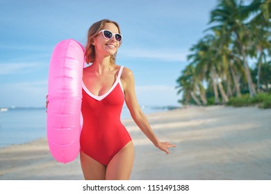 Enjoying suntan and vacation. Outdoor portrait of pretty young woman in red swimsuit with inflatable ring on tropical beach.