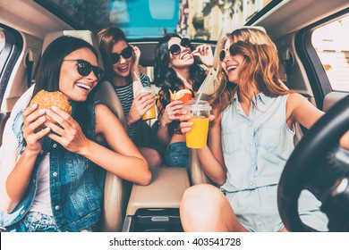 Enjoying Their Lunch In The Car. Four Beautiful Young Cheerful Women Looking At Each Other With Smile And Eating Take Out Food While Sitting In Car