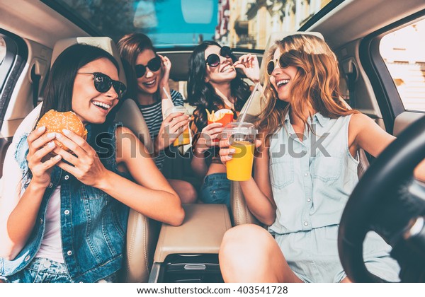 Enjoying their lunch in the car. Four beautiful young\
cheerful women looking at each other with smile and eating take out\
food while sitting in\
car