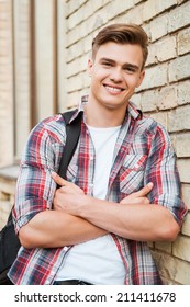 Enjoying Time In College. Handsome Young Man Carrying Backpack On One Shoulder And Smiling While Leaning At The Brick Wall 