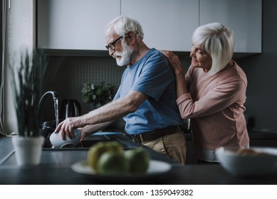 Enjoying time every moment. Waist up side on portrait of senior man washing dishes while his wife tenderly putting hands on his back - Powered by Shutterstock