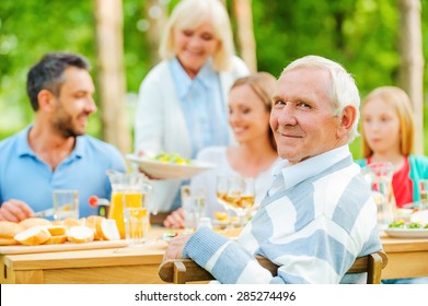 Enjoying time with family. Happy family of five people sitting at the dining table outdoors while senior man looking over shoulder and smiling  - Powered by Shutterstock